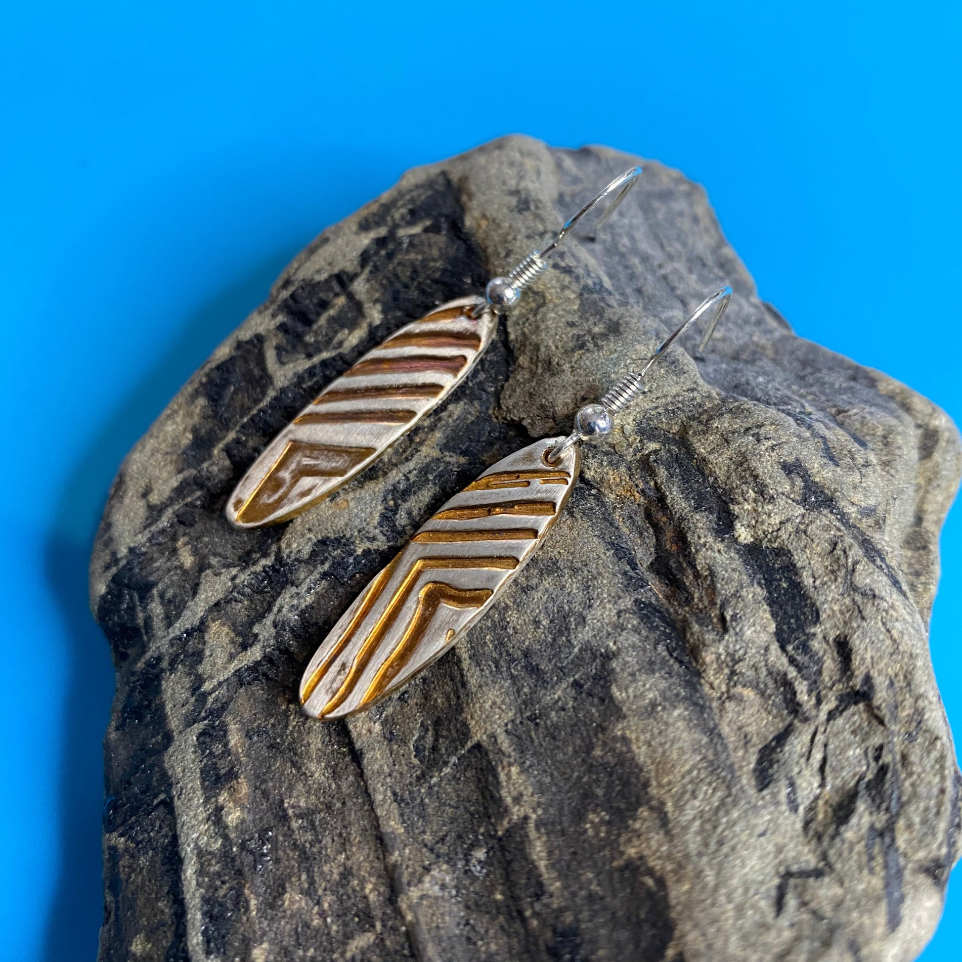 Pair of gold and silver feather-shaped earrings on a textured stone surface with a blue background