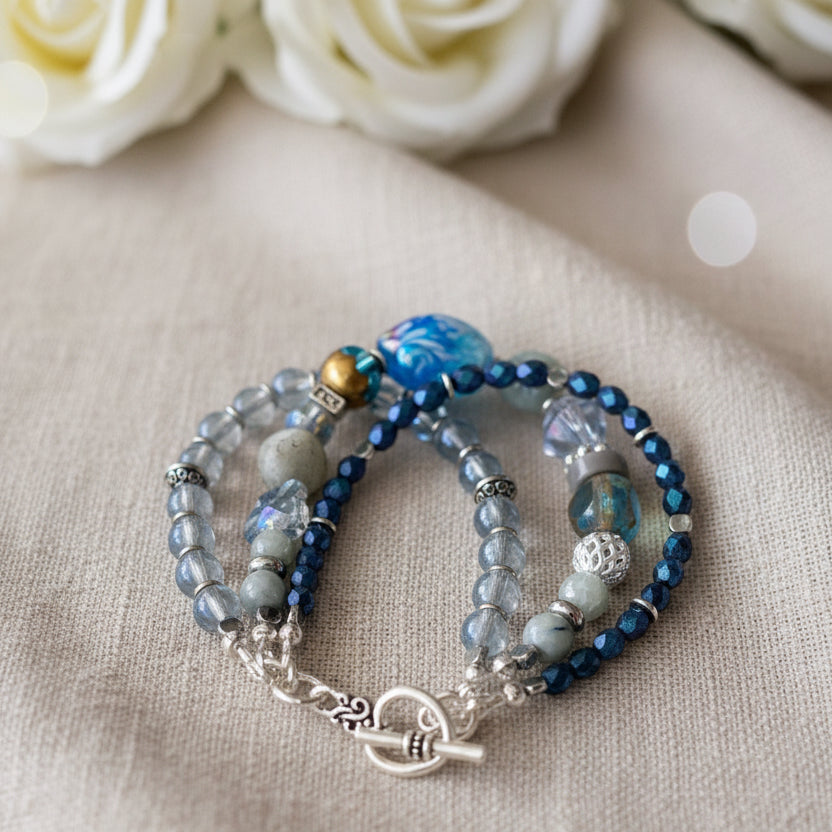 Three strand handmade beaded bracelet on a beige linen cloth with soft white roses in the background. The bracelet is blue with glass bead accents, lamp work and dark blue AB Czech glass.