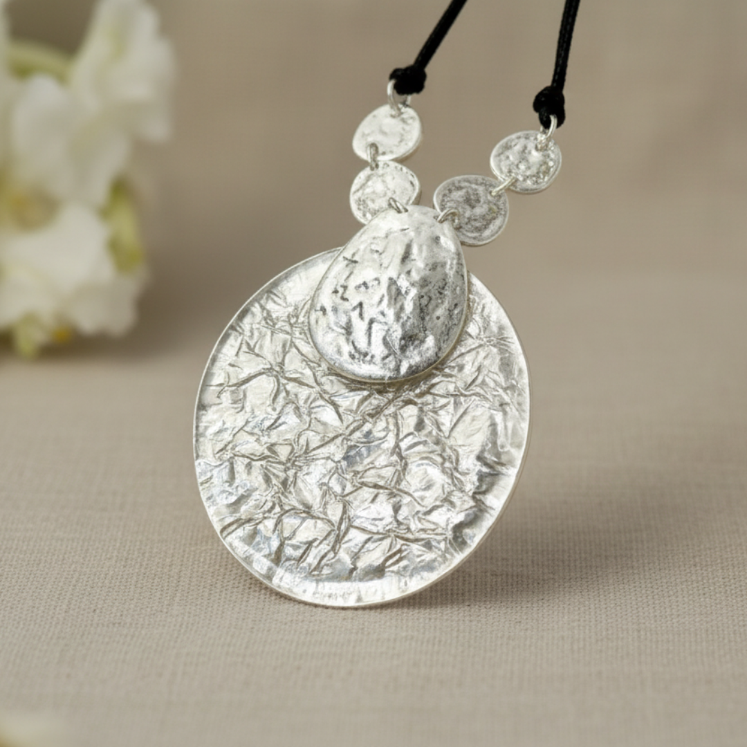 A silver disc fashion necklace with leather accents placed on a light linen fabric, with a floral decoration in the background. Necklace is designed by Jacqueline Kent.