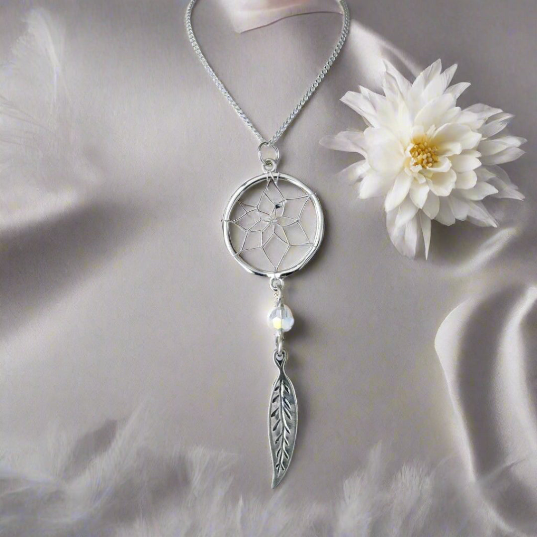 Sterling silver dreamcatcher necklace with feather accent charm on a background of grey satin with a white chrysanthemum.