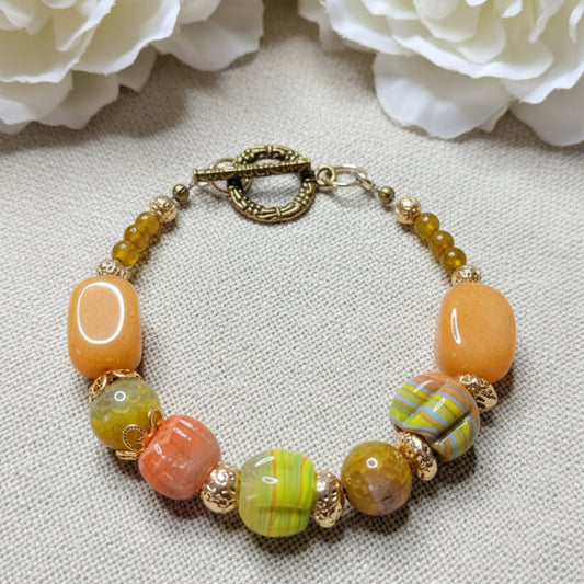 Beaded bracelet with orange, green, and gold beads on a textured linen background.