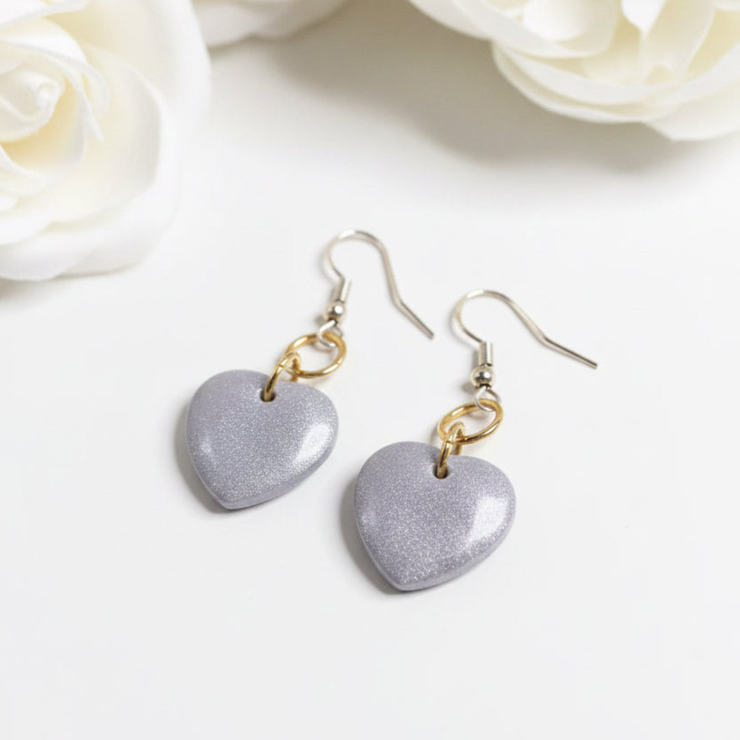 A pair of dangling heart-shaped earrings with a textured, glittery lavender-purple finish and gold-tone hooks, displayed on a solid sage green background.