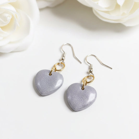 A pair of dangling heart-shaped earrings with a textured, glittery lavender-purple finish and gold-tone hooks, displayed on a solid sage green background.