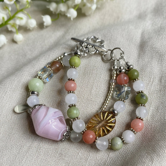 Multicolored beaded bracelet with a pink lamp work focal stone on a light green background.  Handmade in Canada by Arlie Quinn of Arlana Exquisite Styles.