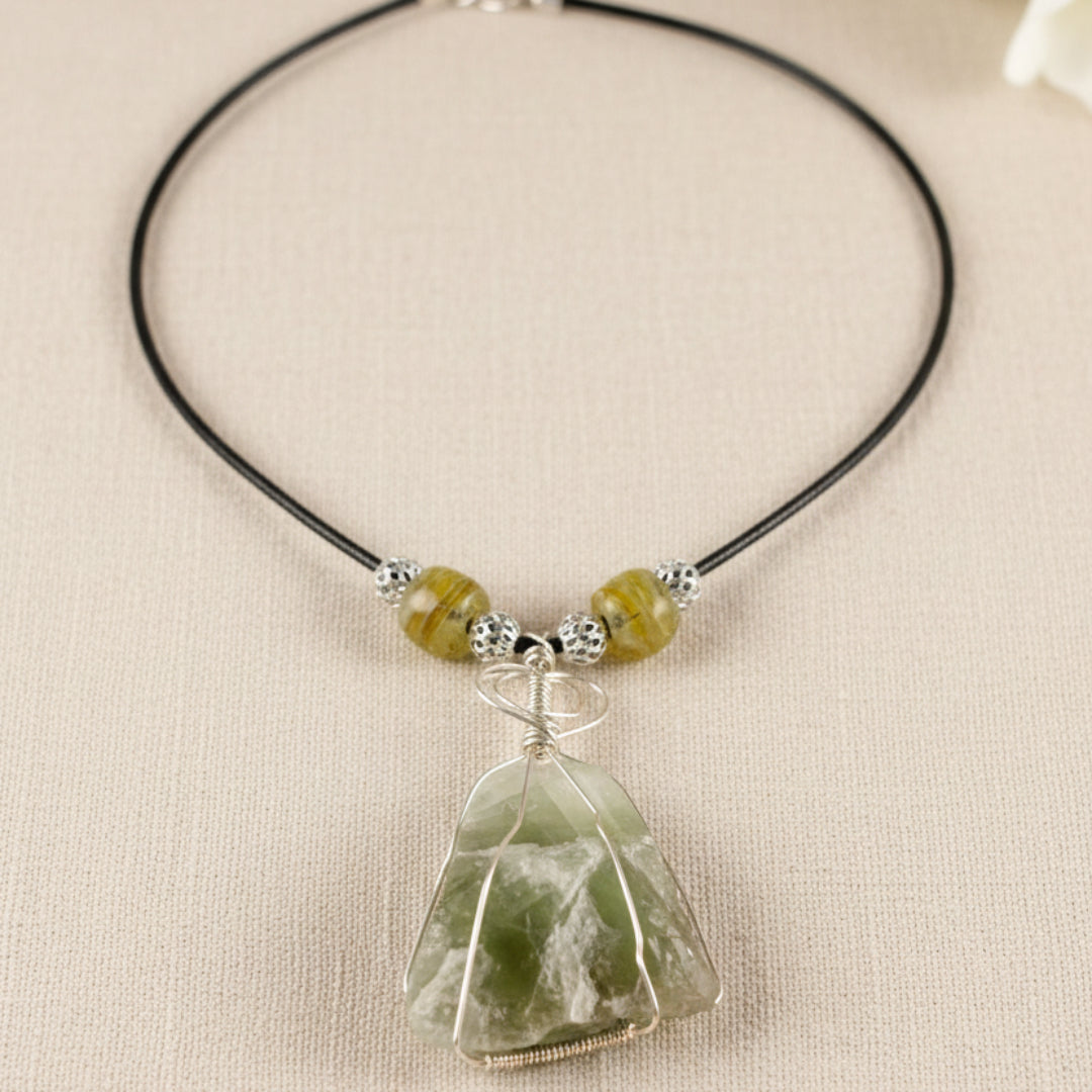 A handmade beaded necklace with a raw green crystal pendant, displayed on a white mannequin bust. The necklace features a black cord, textured silver beads, and translucent olive-green beads, with a large, irregularly shaped green crystal wrapped in silver wire at the center.
