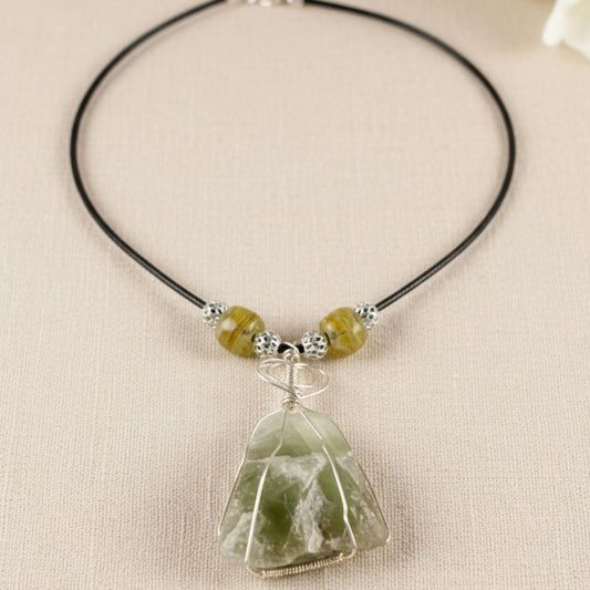 A handmade beaded necklace with a raw green crystal pendant, displayed on a white mannequin bust. The necklace features a black cord, textured silver beads, and translucent olive-green beads, with a large, irregularly shaped green crystal wrapped in silver wire at the center.