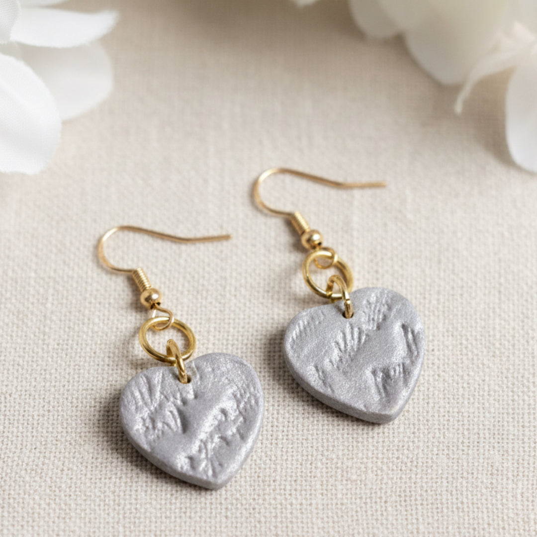 A pair of handmade dangle earrings featuring textured, hammered silver heart-shaped pendants suspended from gold-tone fishhook wires, displayed against a solid linen background.