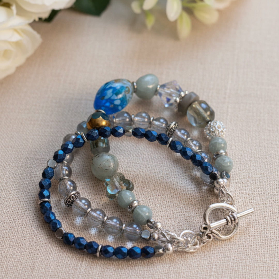 Three strand handmade bracelet on a soft beige linen cloth with white roses in the background.  The bracelet is blue and light grey with silver accents and a silver toggle clasp.